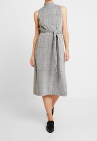 Vestido sin mangas a cuadros grises y negros, con cuello alto, cintura ceñida y longitud midi. Tela suave y textura sutil. Zapatos planos negros.