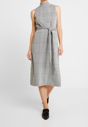 Robe sans manches à carreaux gris et noirs avec col montant, taille ceinturée et longueur midi. Tissu lisse et texture subtile. Chaussures plates noires.