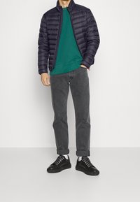 Czarna kurtka puchowa, zielona bluza, szare dżinsy i czarne sportowe buty z sznurowadłami; swobodny strój z różnymi teksturami i kolorami.