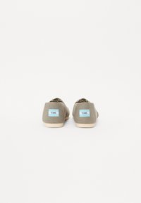 Chaussures slip-on en textile gris avec une semelle claire, présentant une étiquette bleue TOMS au dos. L'intérieur a une doublure à motifs.