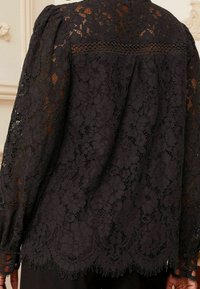 Blusa nera in pizzo con maniche lunghe, motivo floreale e orlo a scallop. Dettagliata con accenti testurizzati e un effetto trasparente sottile.