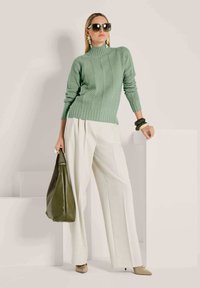 Frau trägt einen grünen Strickpullover und weiße weit geschnittene Hosen, hält eine olivgrüne Tasche und ist mit großen Sonnenbrillen und auffälligen Armbändern accessorized.