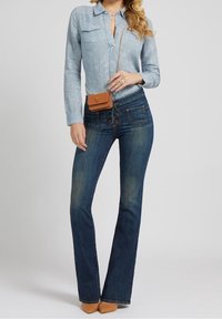 Camisa de botones de color azul claro con un estampado, dos bolsillos en el pecho; combinada con unos jeans acampanados de color azul oscuro y un pequeño bolso cruzado marrón.