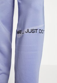 Lila Sporthose mit glatter Textur, elastischem Material und schwarzem, besticktem Text "NIKE_ JUST DO IT" mit silbernem Swoosh-Detail.
