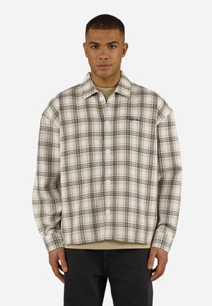 Άνδρας που φοράει πουκάμισο με μπεζ και μαύρο καρό κουμπωτό πάνω από μπλουζάκι ταμπά, με μαύρο παντελόνι, στέκεται με φόντο ένα απλό λευκό τοίχο.