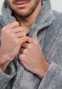 Ropa gris texturizada con un alto cuello y un sutil patrón de espiga, cerrada en el cuello con las manos agarrando las solapas.