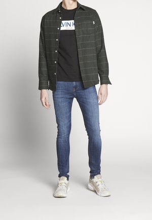 Zielona koszula w kratę zapinana na guziki, noszona na czarnym t-shircie z nadrukiem, do tego niebieskie dżinsy i białe buty sportowe z żółtymi akcentami.