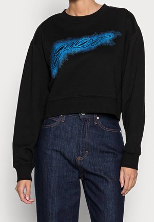 Czarna krótka bluza z długim rękawem z niebieskim logo "guess", zestawiona z ciemnoniebieskimi dżinsami z wysokim stanem na stojącej osobie.