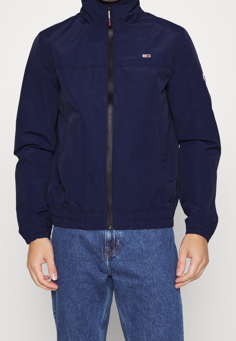 Tmavě modrá bunda s vysokým límcem, předním zipem a elastickými manžetami. Má logo na hrudi a stažený pas.