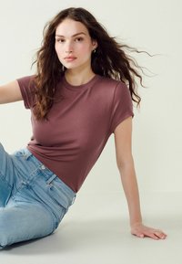 Brun ribbet kortærmet t-shirt med rund hals, parret med lyseblå højtaljede jeans. Modellen sidder med flydende hår.
