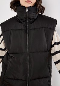 Zwarte puffer vest met hoge kraag, ritssluiting aan de voorkant en zakken. Draagt over een gestreepte trui in crème en zwarte kleuren. Zachte textuur.