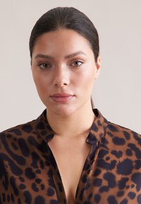 Blusa con stampa leopardata marrone e nera, dal tessuto morbido e trasparente; presenta un colletto e una chiusura con bottoni, sagomata per adattarsi comodamente.
