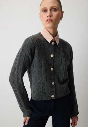 Vrouw draagt een donkergrijze kabelgebreide cardigan met zilveren knopen, perzikkleurig overhemd met kraag en donkere broek tegen een effen achtergrond.