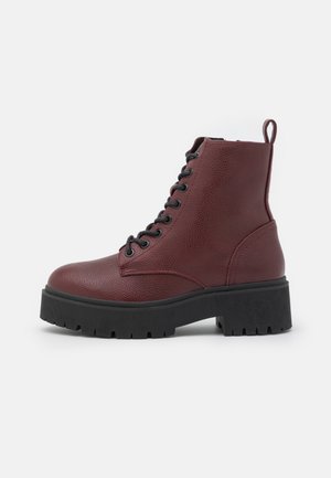 Donkerrode leren enkelboot met veters, getextureerd oppervlak en dikke zwarte geribbelde zool, zijaanzicht op witte achtergrond.