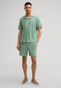 Camicia verde a maniche corte con texture, colletto piatto e bottoni, abbinata a pantaloni short coordinati. Indossata con scarpe slip-on di colore marrone chiaro.
