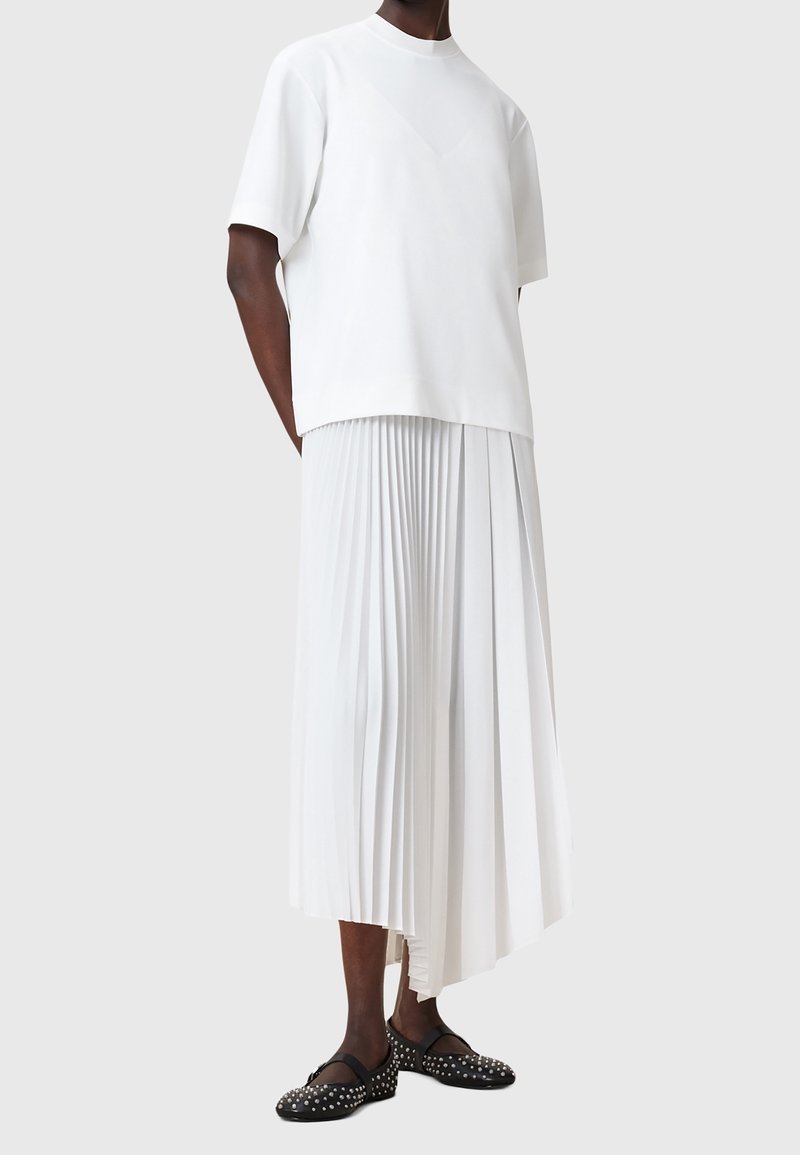 Personne portant un haut blanc oversize à manches courtes et une jupe plissée asymétrique blanche avec des ballerines noires cloutées sur fond gris.