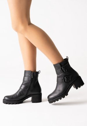 Botas de tobillo de cuero negro con un acabado texturizado, cremallera lateral y acentos de doble hebilla. El tacón grueso y la suela de tacos ofrecen estabilidad.