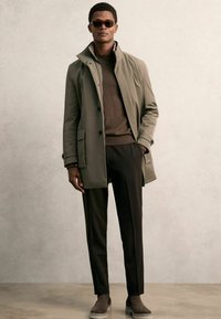Geplanter olivgrüner Mantel mit Stehkragen, schwarzen Knöpfen, zwei Fronttaschen, kombiniert mit einem braunen Pullover, schwarzen Hosen und grauen Slippern.