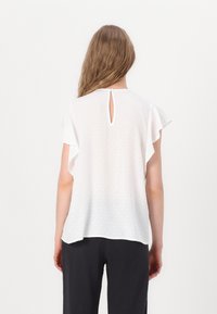 Femme aux longs cheveux portant un chemisier blanc à manches courtes avec des manches volantées et un pantalon noir, vue de dos sur un fond uni.