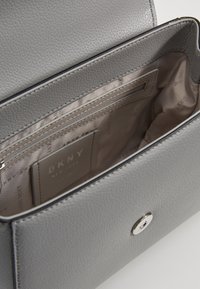 Szara skórzana torba z fakturowaną powierzchnią, wyposażona w kieszeń na suwak i metkę z logo wewnątrz. Zawiera srebrne ozdoby.