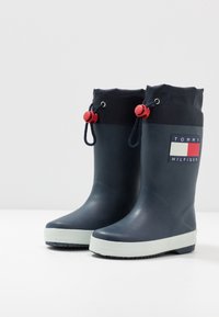 Námornické gumové deštníkové boty s černým textilním manžetou. Mají červené šňůrky, bílou a červenou nášivku s logem a texturovanou bílou podrážku.