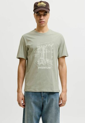 Man draagt een bruine pet en een lichtgroen T-shirt met een witte schets van een caféterras en de tekst "PREMIUM", gecombineerd met een blauwe spijkerbroek.