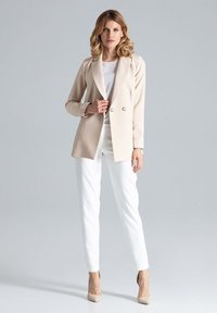 Beige dubbelzijdige blazer met een geknipte revers, gecombineerd met witte slim-fit broeken en lichtbeige hakken, op een grijze achtergrond.