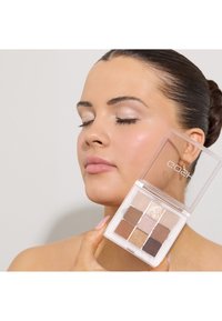 Oogschaduwpalette met negen tinten in een rechthoekige doorzichtige verpakking, met neutrale tinten, gouden accenten en een zachte textuur. In de hand gehouden.