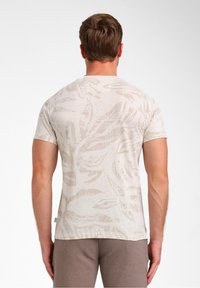 T-shirt beige clair avec un motif de feuilles subtil, manches courtes, col rond et une texture douce. Associé à un pantalon beige foncé.