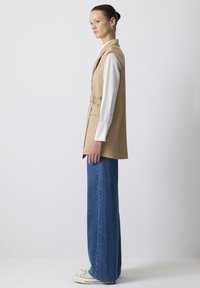 Beige mouwloze vest met een cinched taille, zijzakken en kraag over een wit overhemd met lange mouwen, gecombineerd met wijde blauwe denim broek.