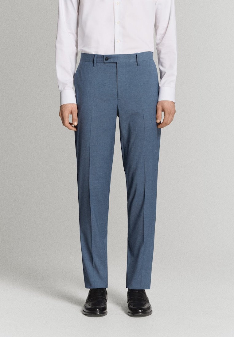 Muž oblečený v prispôsobených modrých nohaviciach na spoločenské príležitosti, bielej košeli na gombíky a čiernych kožených spoločenských topánkach stojaci pred jednoduchým pozadím.