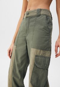 Olivově zelené cargo kalhoty s uvolněným střihem, vybavené knoflíkovým zapínáním, elastickým páskem a velkou boční kapsou na stehně.