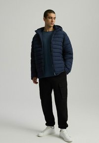 Granatowa kurtka puchowa z poziomym pikowaniem, z odpinanym kapturem, w połączeniu z niebieskim swetrem, czarnymi spodniami cargo i białymi trampkami.