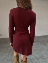 Bordowa sukienka do kolan o dopasowanym fasonie, z długimi rękawami i delikatnym zakładanym detalem w talii; noszona z brązowymi botkami za kostkę.