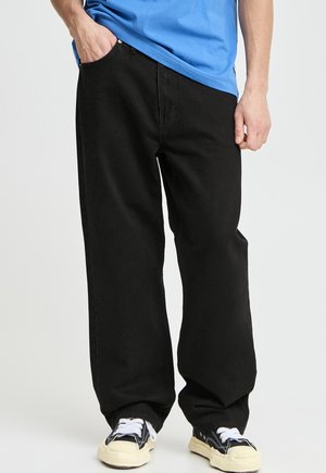 Person iført sorte løstsiddende jeans, blå skjorte og sort-hvide sneakers, stående med den ene hånd i lommen.