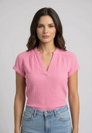 Femme aux cheveux mi-longs et foncés, portant un chemisier rose à manches courtes avec col en V et un jean bleu clair, face à l’avant avec une expression neutre.