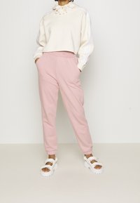 Krämfärgad kort hoodie med dragsko, matchande rosa tapered byxor, vita chunkysandaler, mjukt tyg och subtil branding på plaggen.