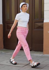 Weißes kurzärmeliges Oberteil, rosa Hose und weiße Sandalen mit Schnalle. Hat einen Bucket Hat, Tattoos und präsentiert eine Geh-Pose vor einem einfarbigen Hintergrund.