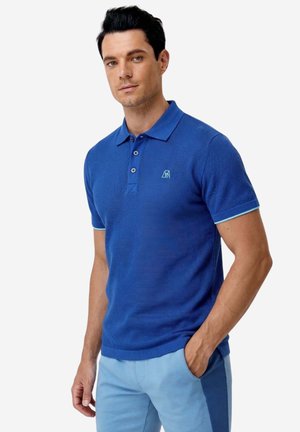 Uomo in piedi che indossa una polo blu reale strutturata con un piccolo logo e pantaloni azzurri, mano in tasca, su sfondo semplice.