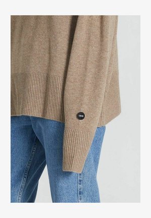 Rudas megztinis su raukšlėta tekstūra ir rankogaliais, su šoniniu etiketu, ant kurio užrašyta "IAYS". Derinamas su šviesiai mėlynomis džinsinėmis kelnėmis.