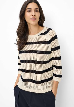 Junge Frau mit langen dunklen Haaren, die einen cremefarbenen, gestrickten Pullover mit schwarzen horizontalen Streifen und dunkle Hosen trägt, mit den Händen in den Taschen stehend.