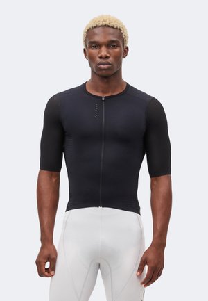 Personne athlétique aux cheveux blonds portant un maillot de cyclisme noir moulant à manches courtes et un cuissard blanc rembourré, face vers l’avant.