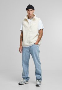 Gilet en polaire crème avec un col haut, fermeture éclair et poche avant, associé à un jean bleu clair et des baskets de sport.