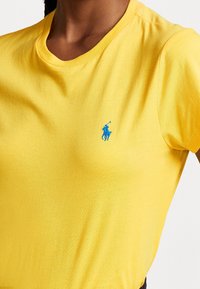 Detailní záběr osoby na sobě má jasně žluté tričko s malým logem polo hráče modré barvy na hrudi.