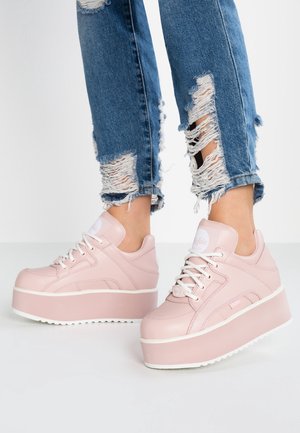 Pink platform sneakers with white laces worn with blue ripped skinny jeans against a plain white background.