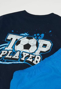 Námořnické tričko s bílým a modrým textem "TOP PLAYER" a grafikou fotbalového míče, v kombinaci s modrými šortkami. Oba kusy jsou vyrobeny z měkké bavlny.