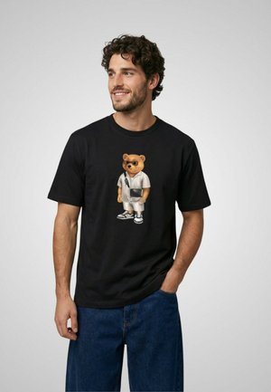 Uomo che indossa una T-shirt nera con grafica di un orso in abbigliamento bianco, borsa a tracolla e occhiali da sole, abbinata a jeans blu, in piedi davanti a uno sfondo semplice.