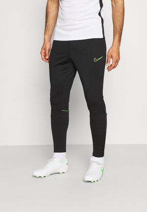 Mann trägt schwarze Nike-Trainingshose mit grünem Logo, weiße Sportsocken, weiße Fußballschuhe mit grünen Akzenten und ein weißes Hemd.