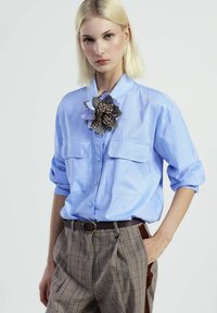 Camicia azzurro chiaro con bottoni e due tasche sul petto, abbinata a pantaloni a quadri marroni e un grande accento floreale multi-texturato al colletto.