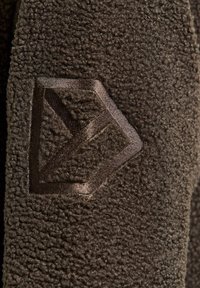Gros plan d'un tissu texturé brun foncé avec un emblème géométrique en forme de flèche cousu sur la surface.